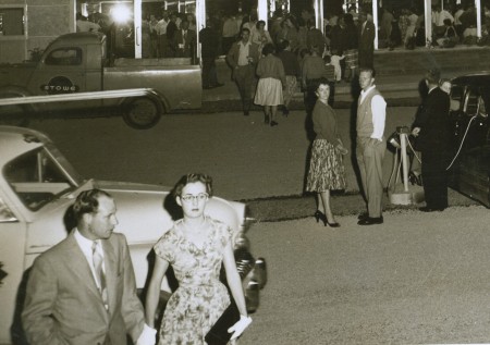 A close-up of the first image showing the semi-formal attire of the couples. The men are wearing ties and jackets and the women are wearing dresses with jewellery and delicate gloves, and carrying clutches.